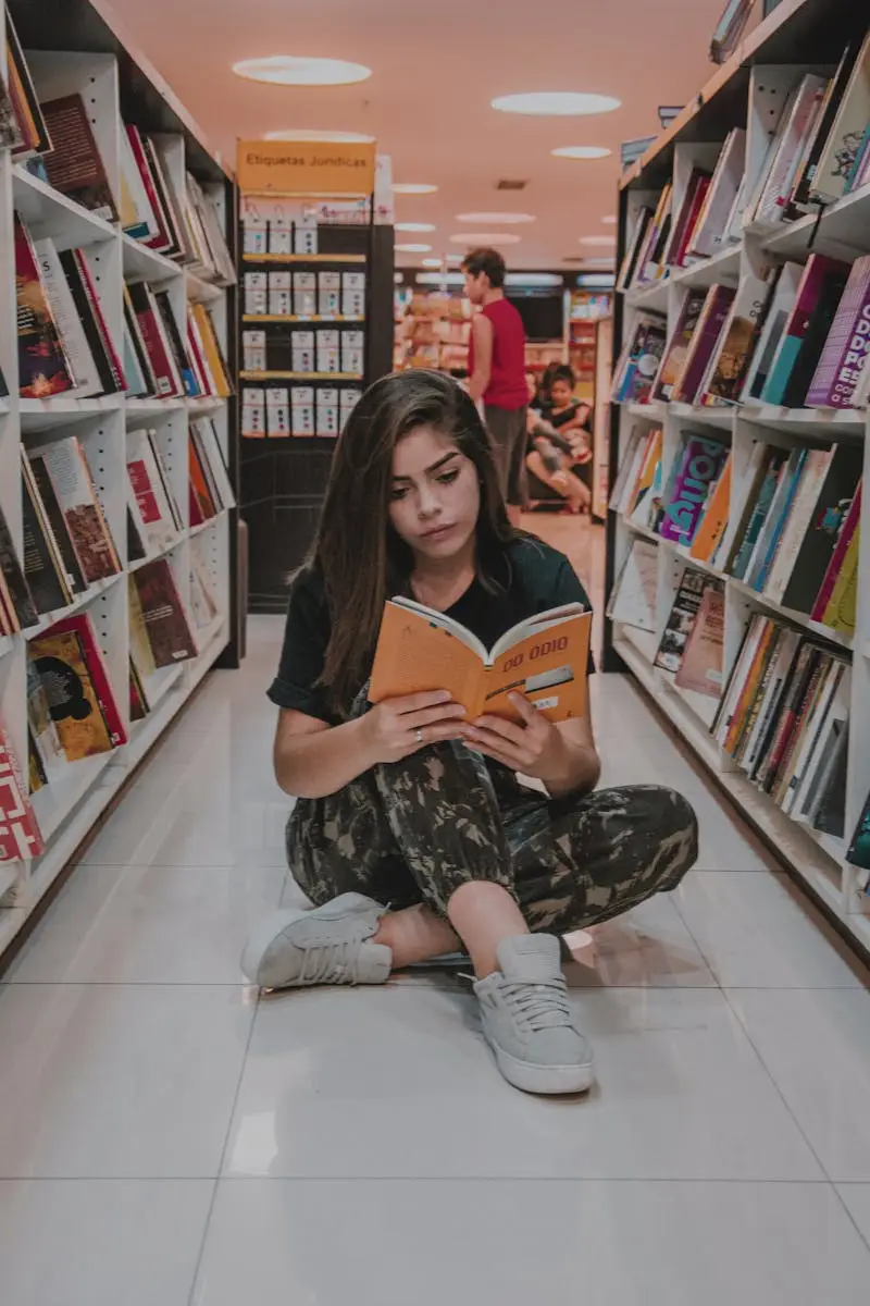 Tentang Kami Young woman sitting on the floor and reading a book in a modern bookstore with shelves full of colorful books.